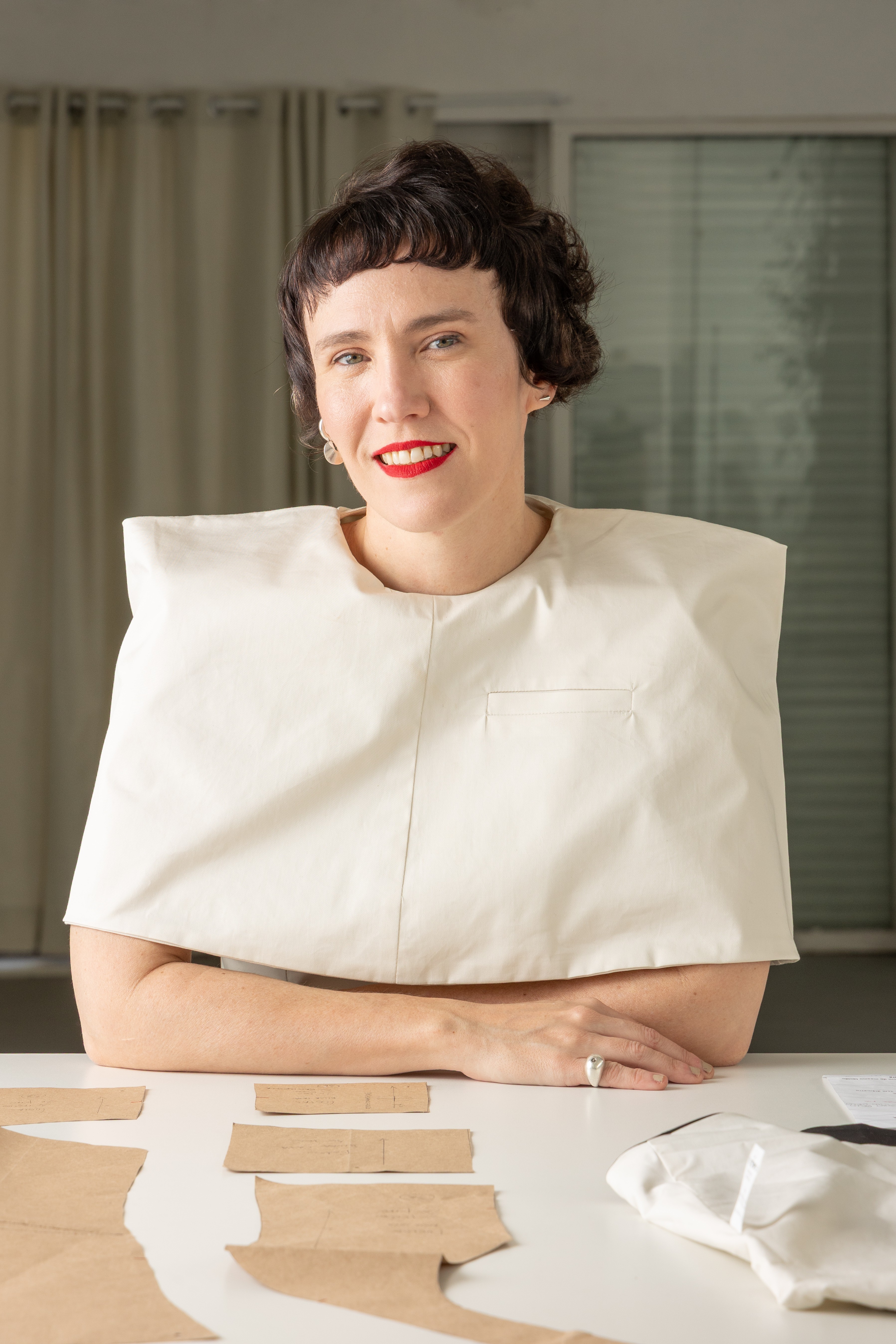 Mulher de cabelo curto e escuro, com franja, sorri para a c&acirc;mera. Veste um colete creme com ombros estruturados e um bolso frontal. Ela est&aacute; sentada em uma mesa branca com moldes de papel pardo espalhados e um tecido branco dobrado. Ao fundo, cortinas claras e uma janela com persianas.