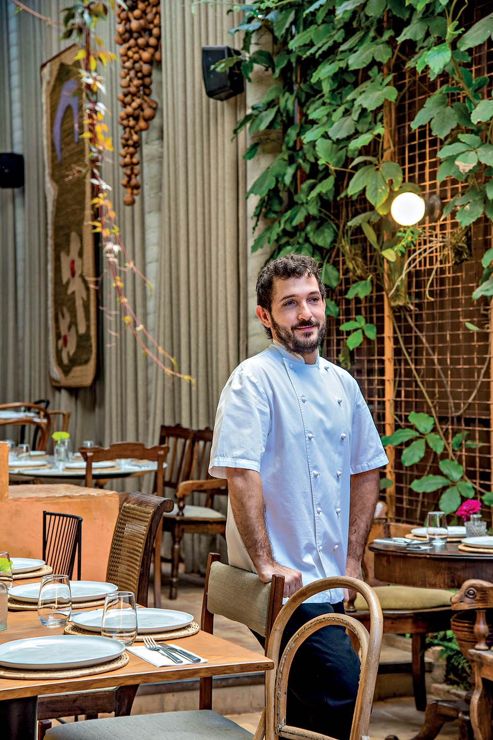 Homem barbudo de jaleco branco, sorrindo e apoiado em uma cadeira de madeira, em um restaurante com mesas postas e plantas trepadeiras na parede