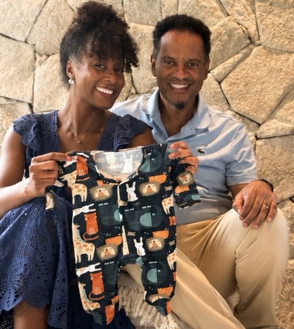 Casal sorridente, mulher de vestido azul e homem de camisa polo clara, segurando um macacão de bebê com estampa de animais da floresta, como girafas e leões, em frente a uma parede de pedras