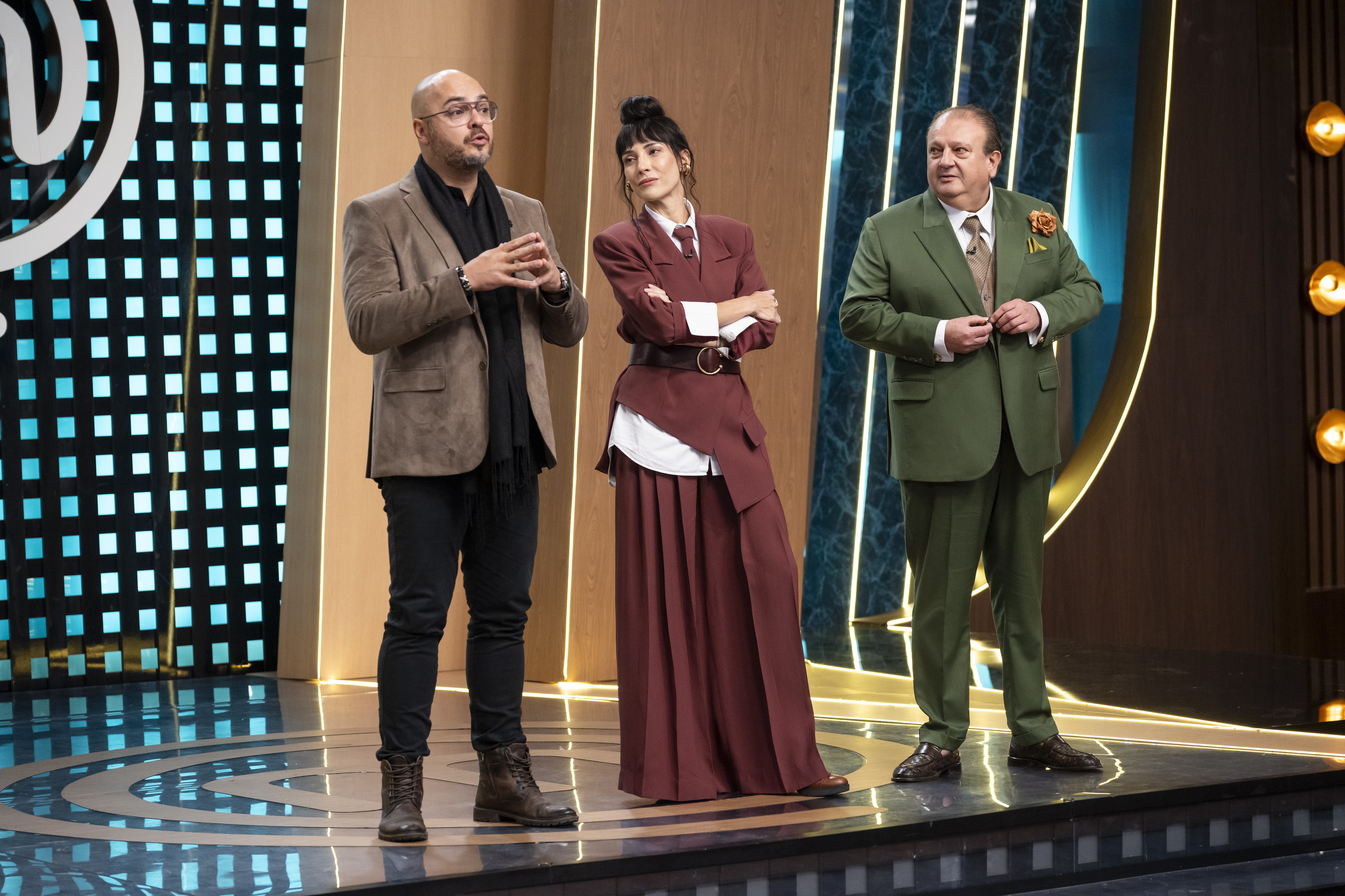 Três jurados, dois homens e uma mulher, em um palco de programa de culinária. O homem à esquerda, careca e de óculos, veste blazer marrom e cachecol preto, gesticulando. A mulher ao centro usa um conjunto vinho com saia longa e blusa branca, braços cruzados. O homem à direita, de terno verde-oliva, abotoa o paletó. O palco tem iluminação azul e dourada