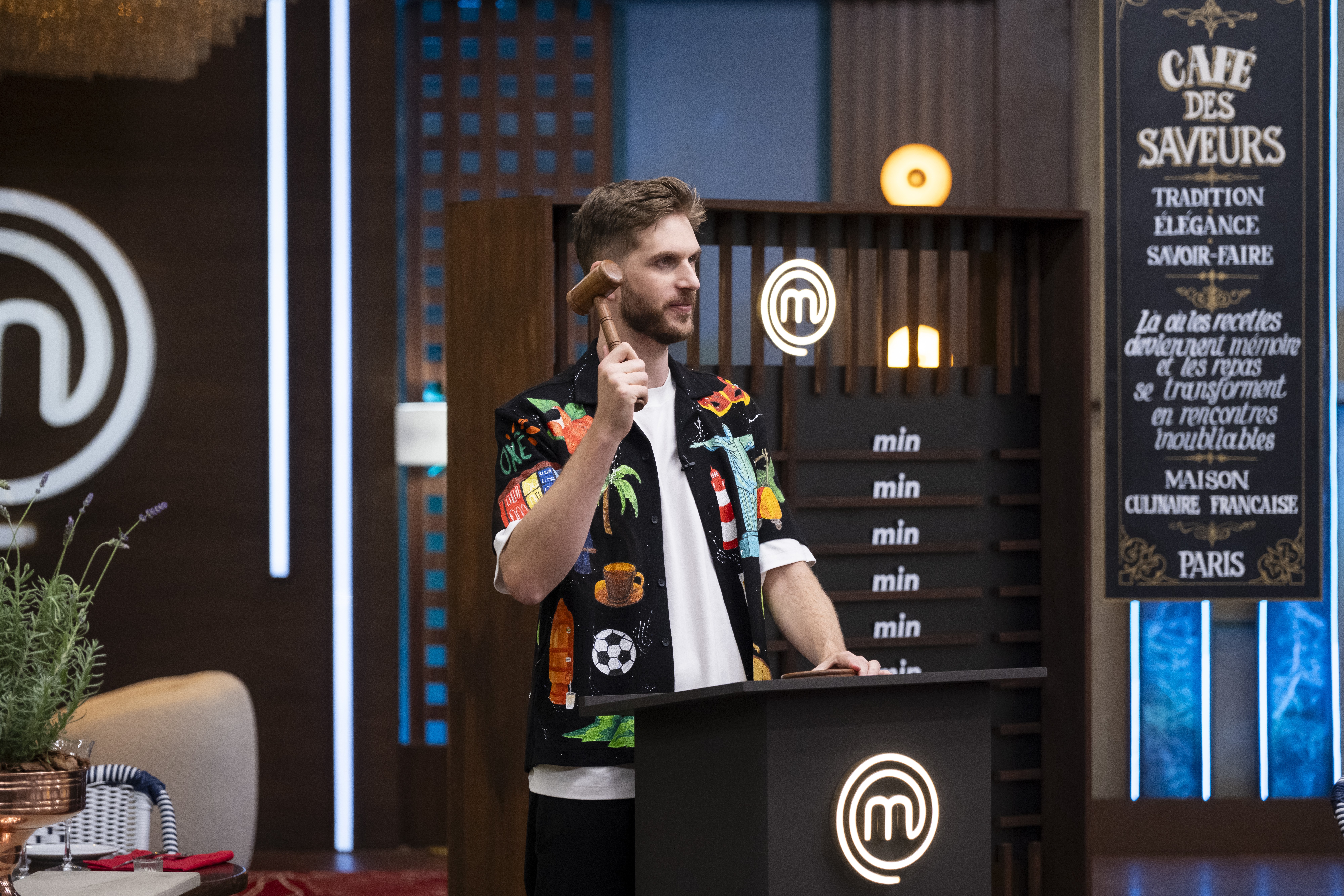 Homem barbudo de camisa estampada colorida e camiseta branca, segurando um martelo de madeira, em pé atrás de um púlpito preto com o logo M do MasterChef. Ao fundo, um painel com min repetido e uma placa preta com Café des Saveurs em francês