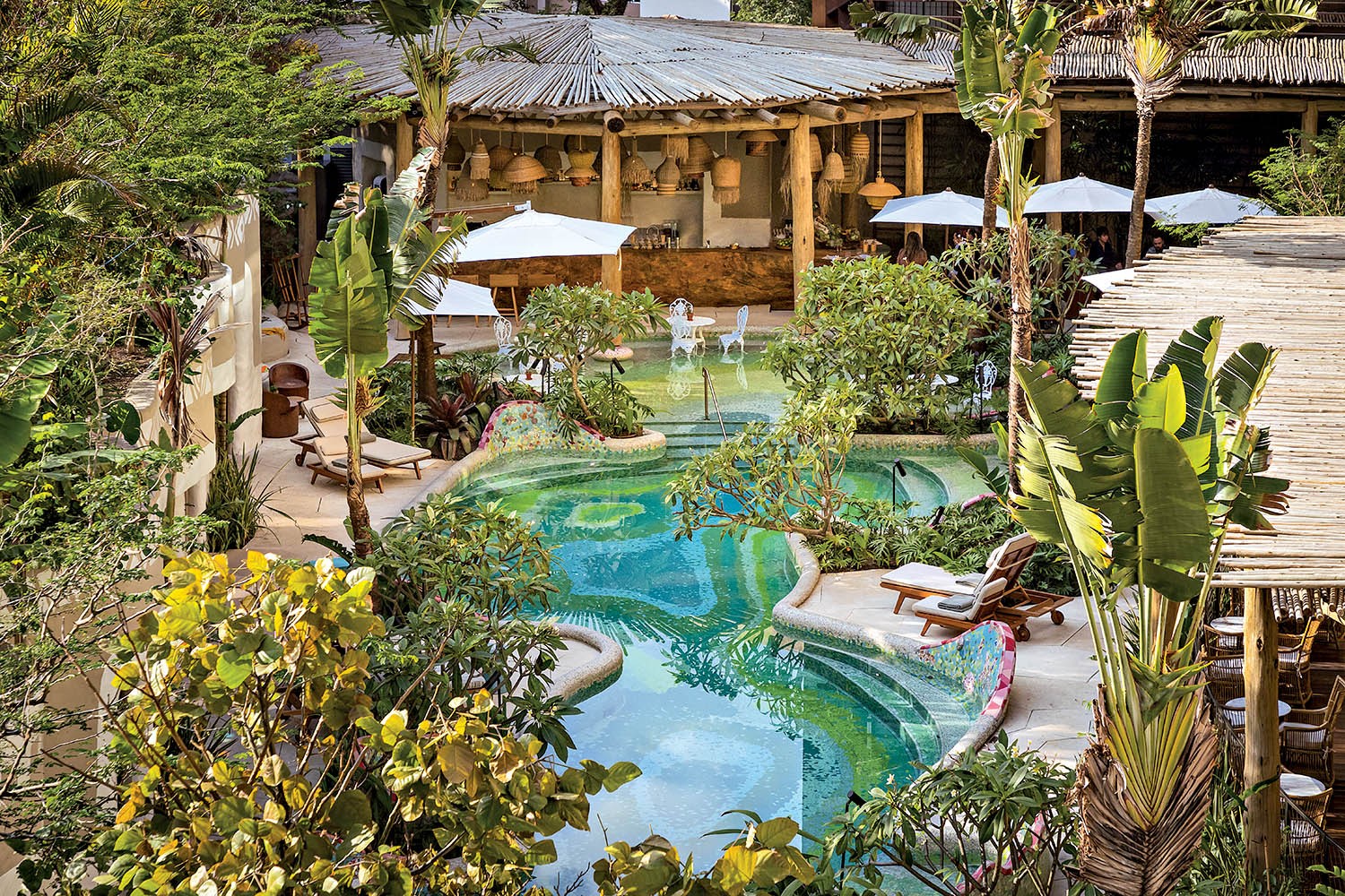 Vista a&eacute;rea de um resort tropical com piscina de formato org&acirc;nico em tons de azul e verde, cercada por espregui&ccedil;adeiras e vegeta&ccedil;&atilde;o exuberante. Ao fundo, um bar r&uacute;stico com telhado de palha e lumin&aacute;rias penduradas, e mesas com guarda-s&oacute;is brancos. O ambiente &eacute; de relaxamento e natureza