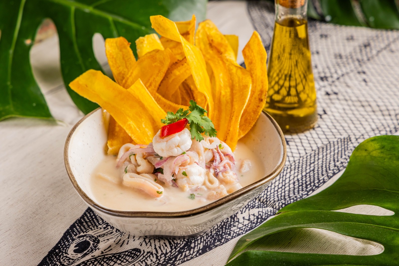 Ceviche de camarão e lula em molho cremoso, guarnecido com coentro e pimenta vermelha, acompanhado de chips de banana-da-terra crocantes. Uma garrafa de azeite e folhas verdes tropicais decoram a mesa com toalha estampada