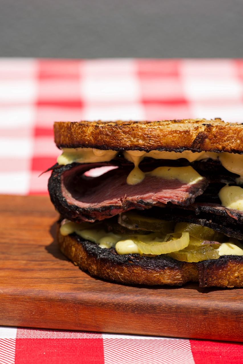 Sanduíche de pastrami com fatias de carne vermelha, picles e molho cremoso, servido em pão torrado sobre uma tábua de madeira, com toalha xadrez vermelha e branca ao fundo