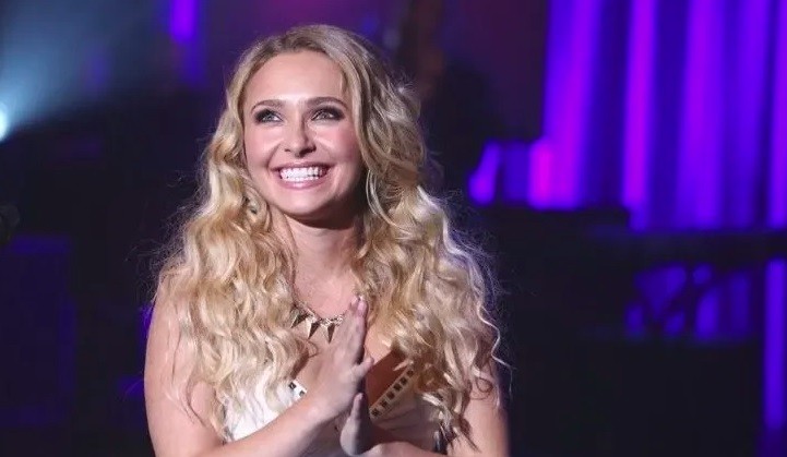 Mulher loira de cabelos ondulados, sorrindo amplamente com a mão direita sobre o peito, usando um colar de pingentes triangulares, em um palco com iluminação roxa