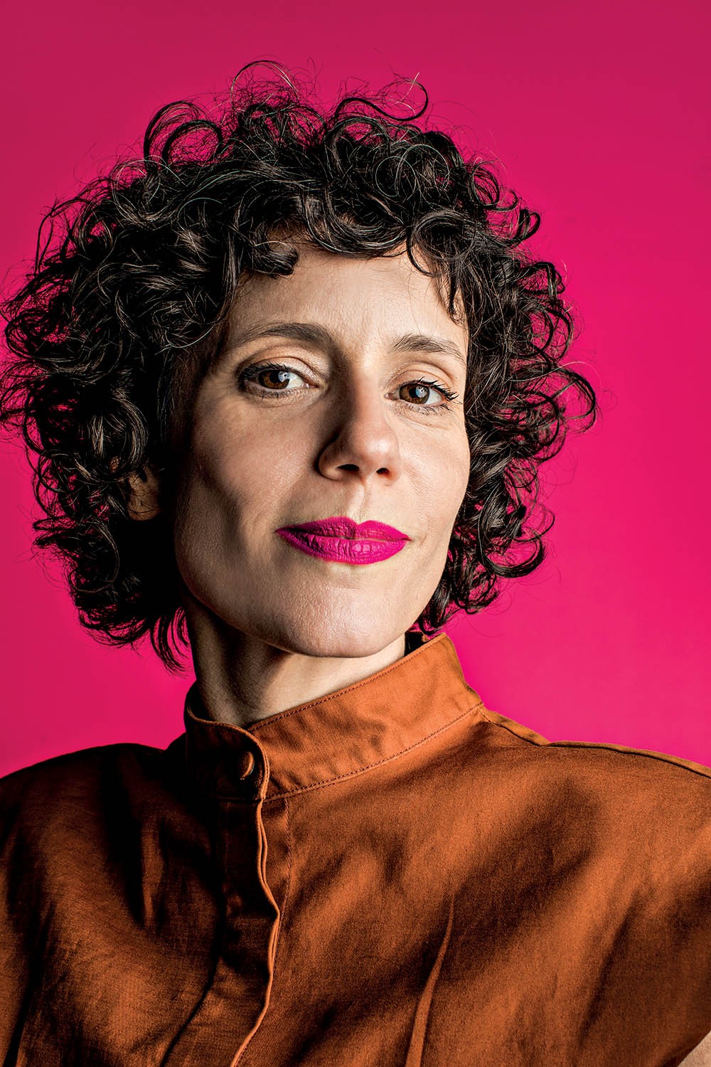 Mulher de pele clara, cabelos curtos e cacheados, vestindo camisa marrom, com batom rosa vibrante, olhando para frente em fundo rosa choque