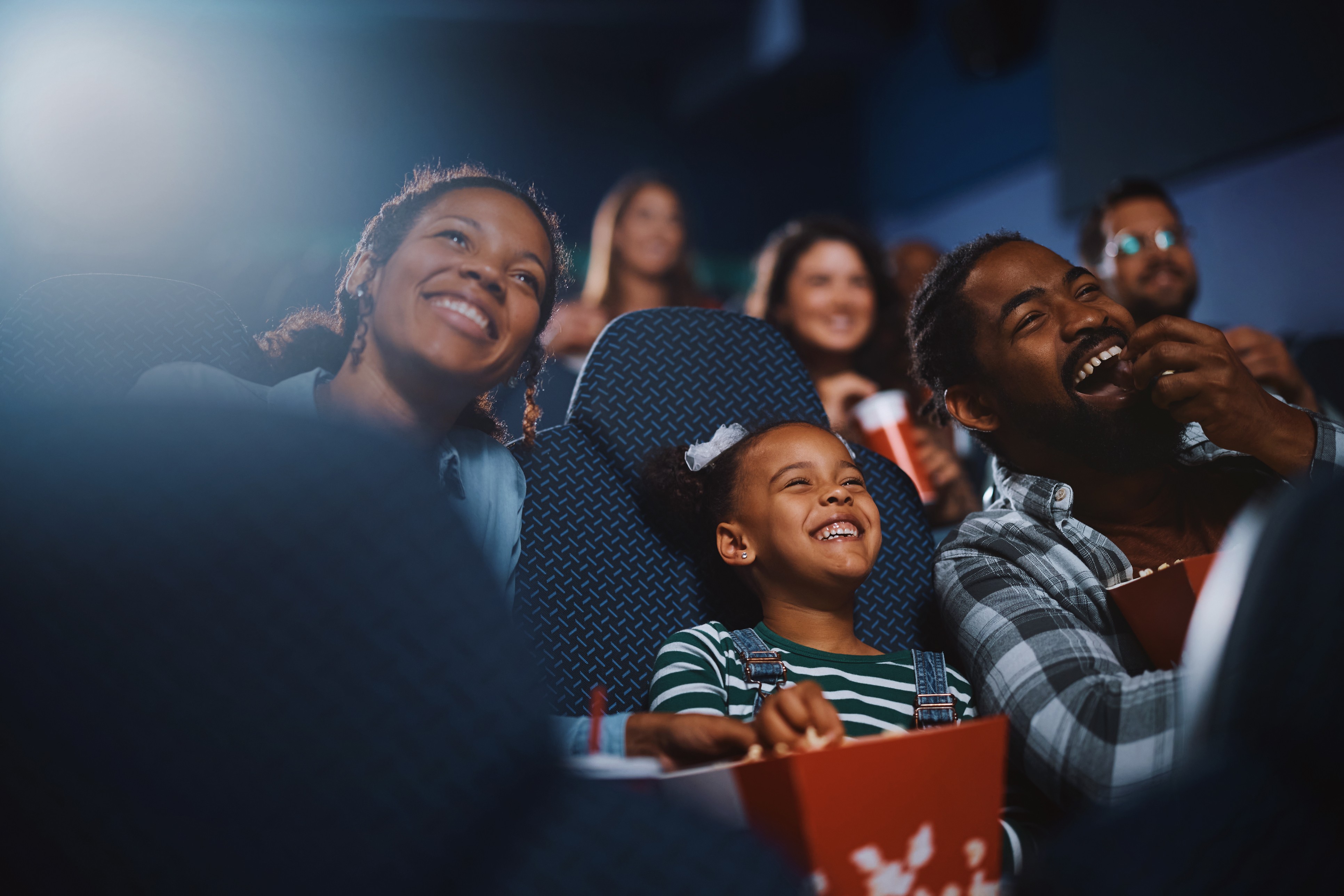 Fam&iacute;lia feliz assistindo a um filme no cinema. Uma mulher, uma menina e um homem negros riem, comendo pipoca.