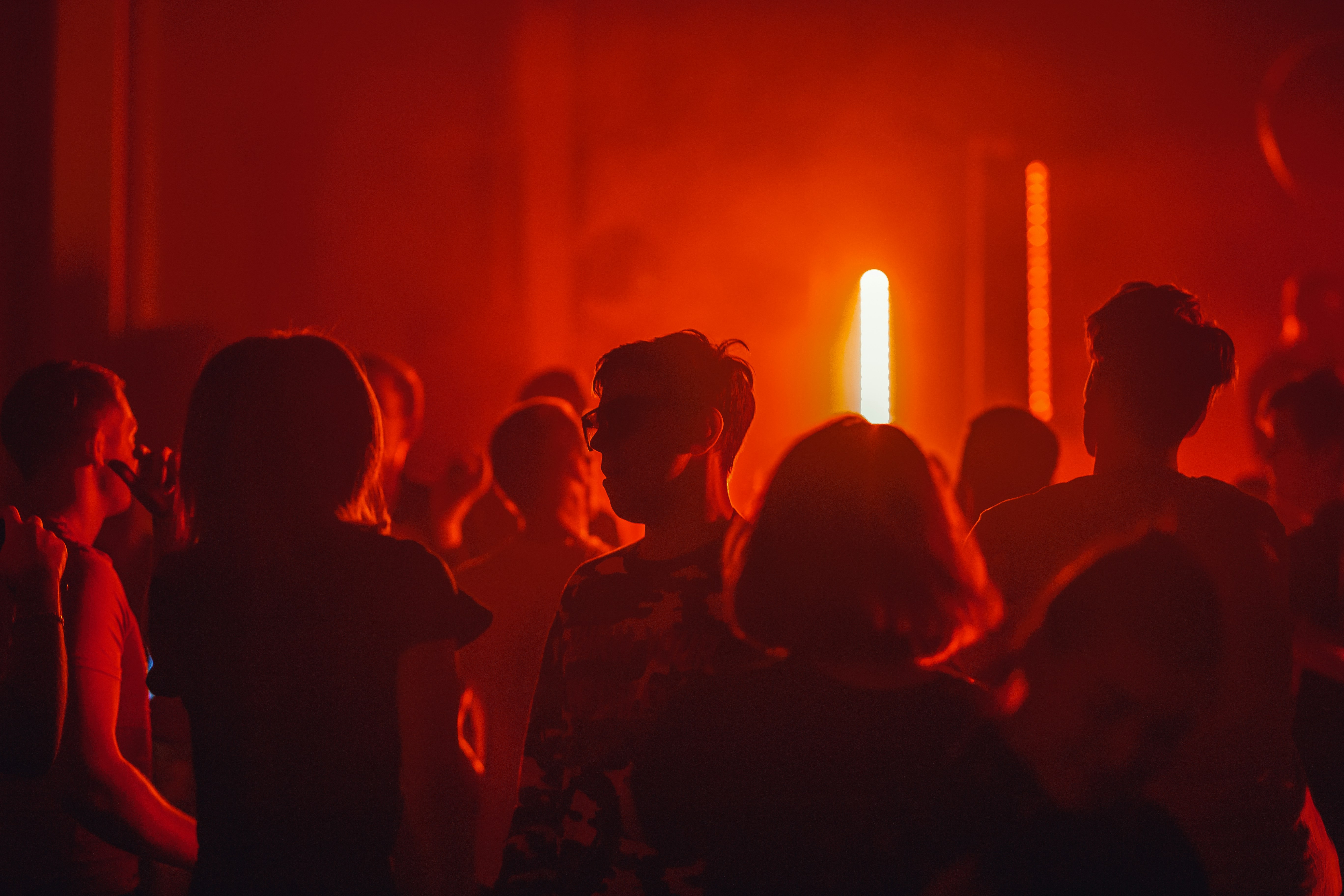 Pessoas em silhueta, iluminadas por luzes vermelhas e uma barra vertical amarela brilhante ao fundo, em um ambiente de festa ou show