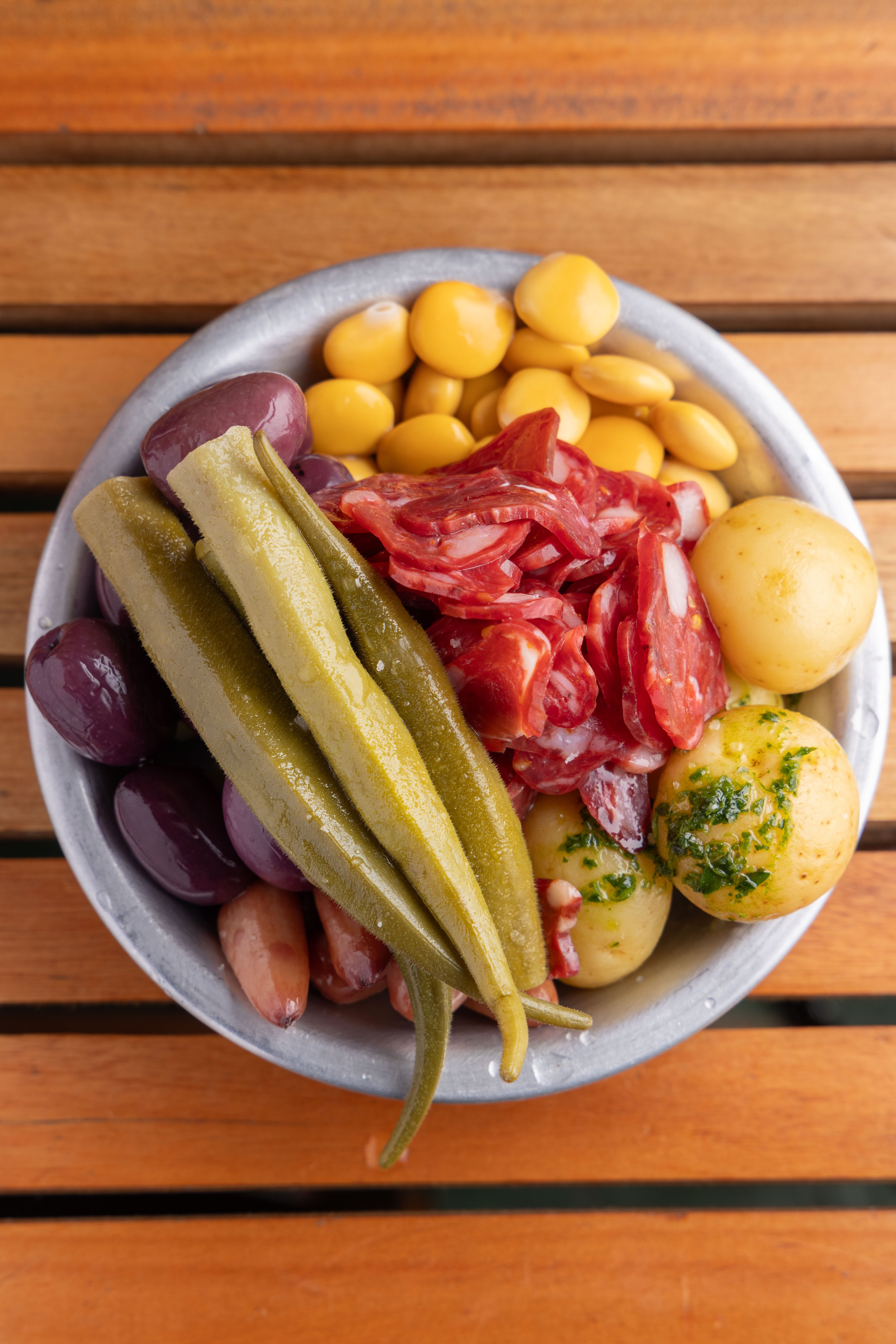 Uma tigela de metal com petiscos variados: tremo&ccedil;os amarelos, rodelas de salame vermelho, azeitonas roxas e marrons, quiabos verdes e batatas cozidas com salsa, sobre uma mesa de madeira