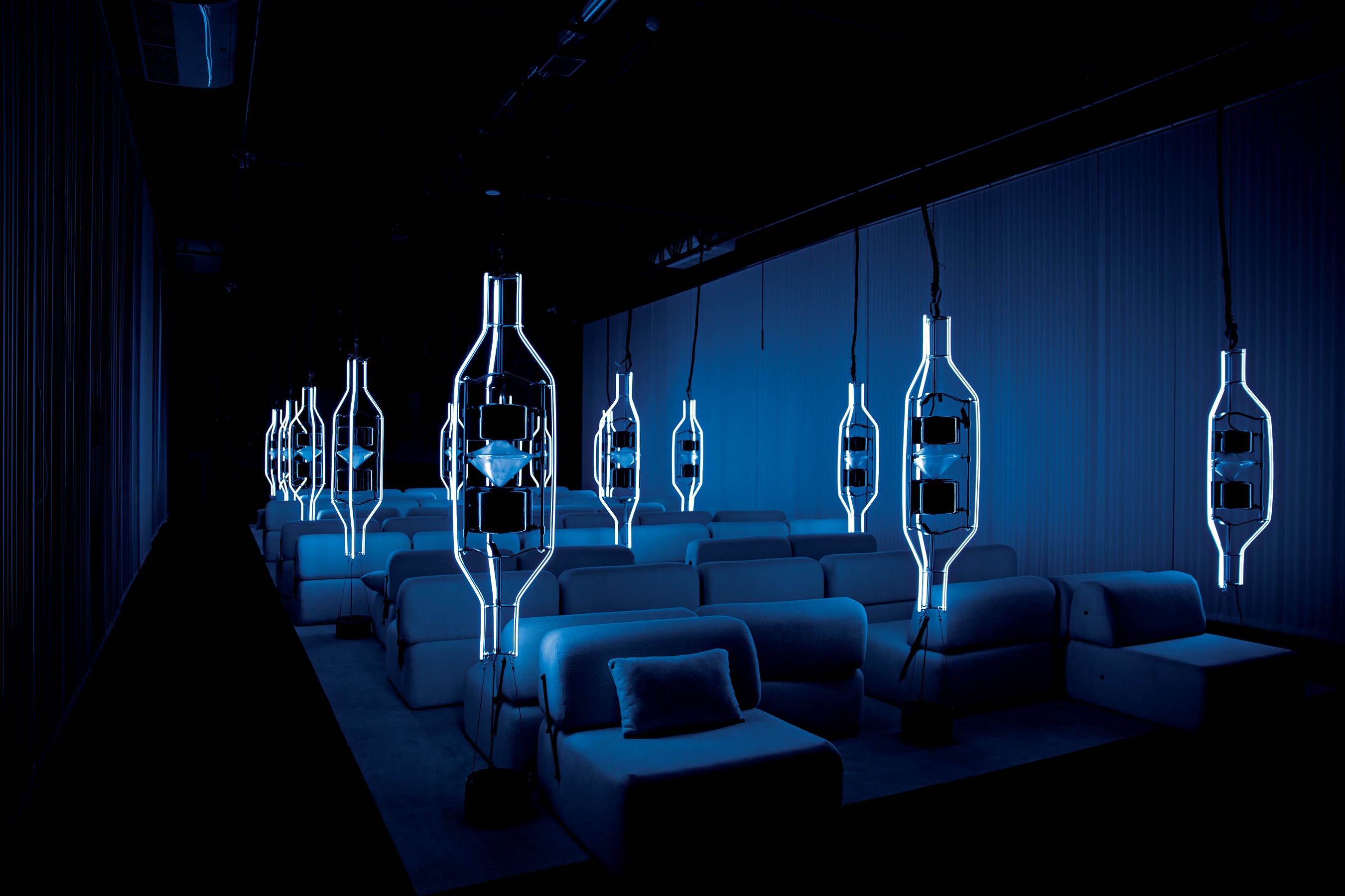 Ambiente escuro com fileiras de poltronas azuis e luminárias penduradas em formato de garrafa, emitindo luz azul neon