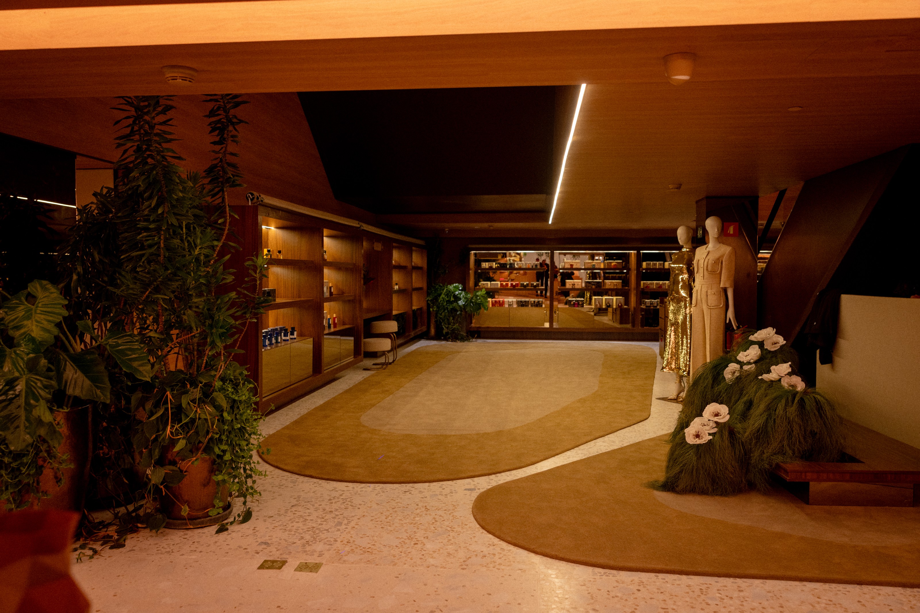 Interior de uma loja ou galeria com iluminação quente, piso claro e tapetes marrons. À esquerda, plantas grandes em vasos. Ao fundo, prateleiras de madeira com produtos e, à direita, dois manequins exibindo roupas, um dourado e outro bege, próximos a uma escada escura e um arranjo floral com flores brancas