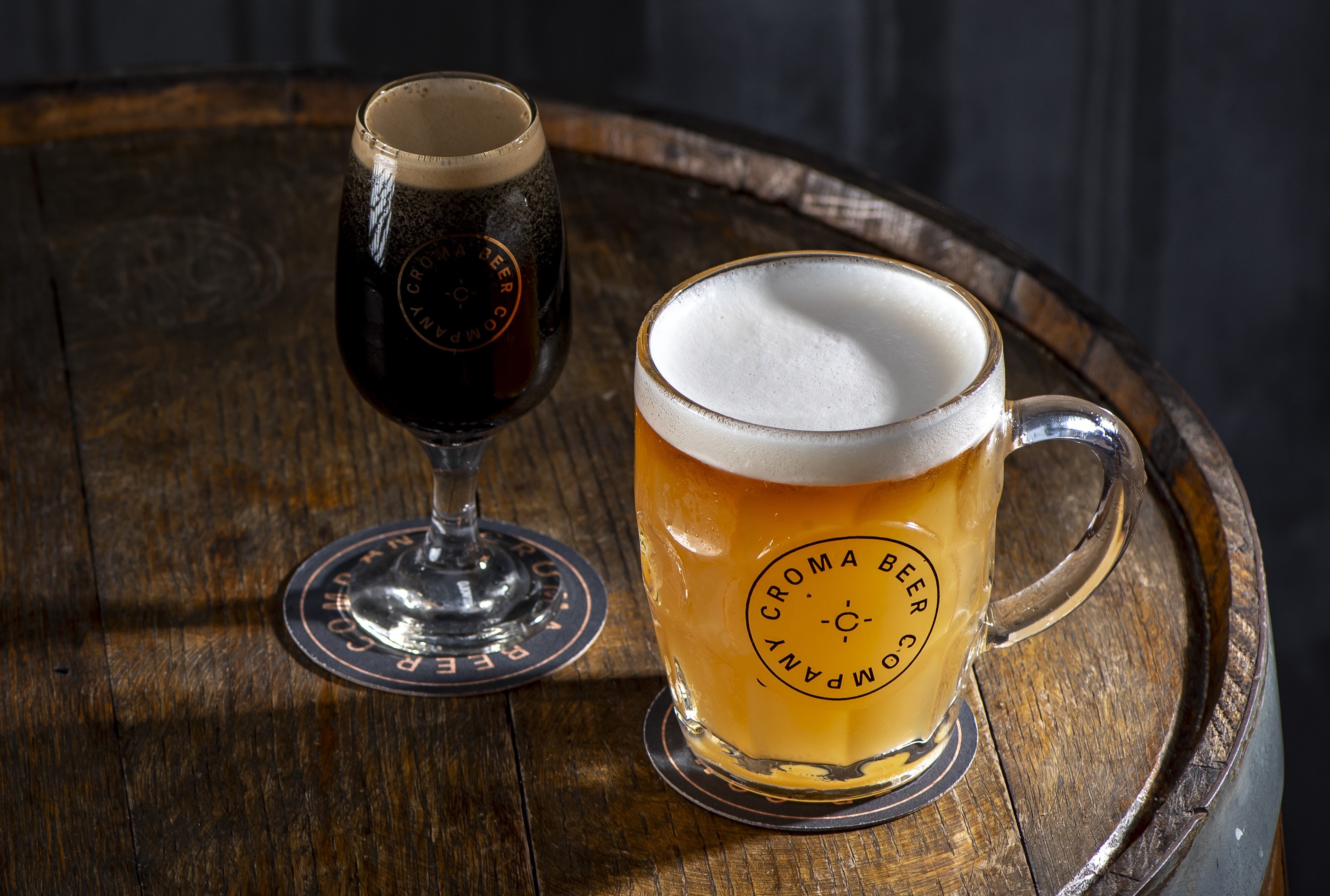 Um copo de cerveja escura e uma caneca de cerveja clara com espuma branca, ambas com o logo Croma Beer Company, sobre um barril de madeira r&uacute;stico