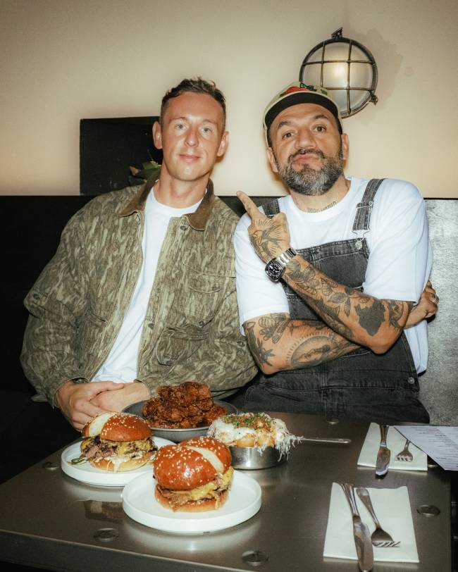 Dois homens sorridentes sentados &agrave; mesa de um restaurante, com v&aacute;rios pratos de comida &agrave; frente. O da esquerda veste jaqueta camuflada e camiseta branca. O da direita, com barba e bon&eacute;, usa macac&atilde;o jeans e camiseta branca, apontando para cima com o dedo indicador. Na mesa, h&aacute; dois hamb&uacute;rgueres, frango frito e um prato com queijo derretido
