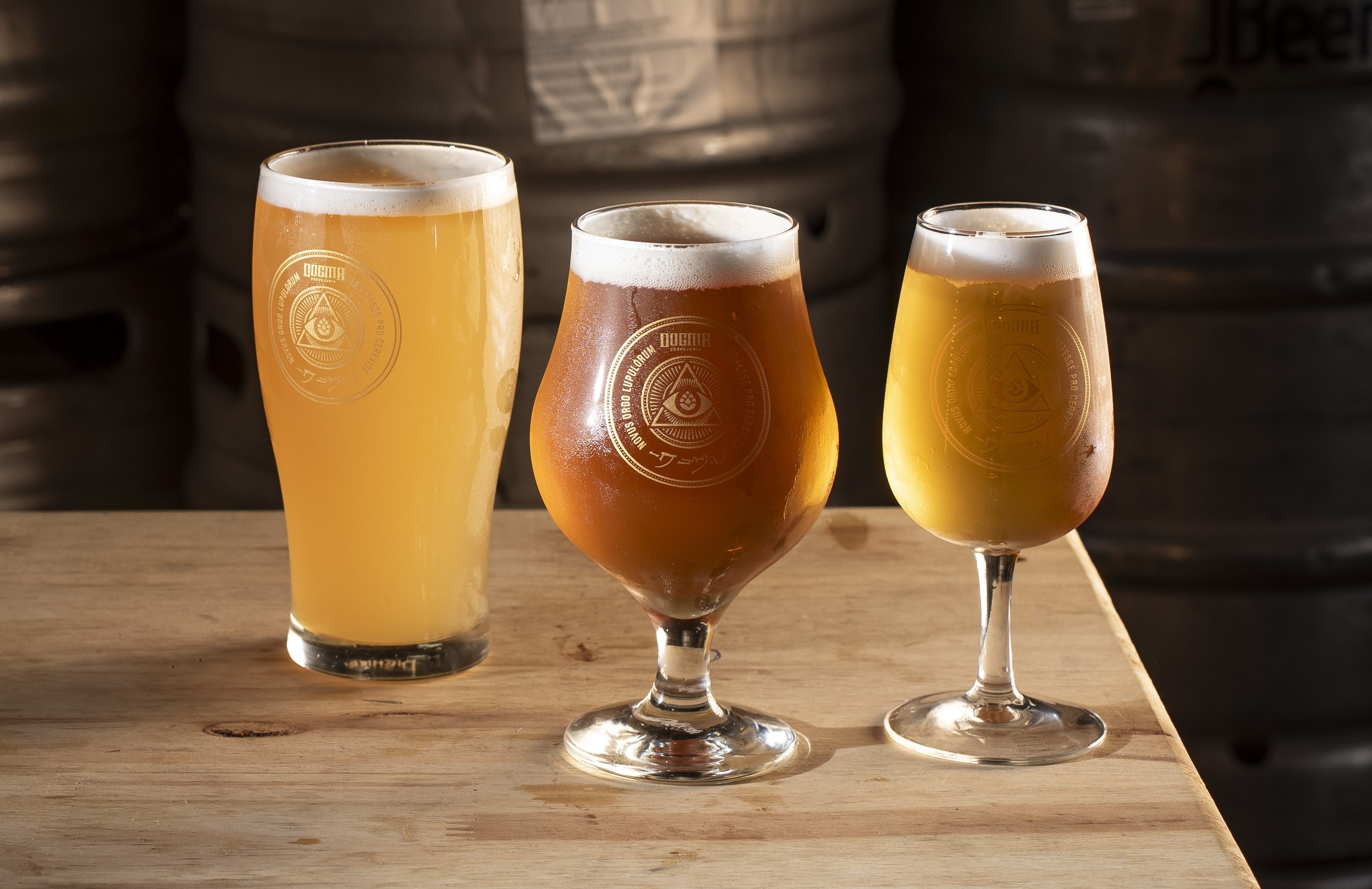 Tr&ecirc;s copos de cerveja com espuma branca, de cores e formatos diferentes, sobre uma mesa de madeira clara. O primeiro &eacute; um pint de cerveja clara, o segundo &eacute; uma ta&ccedil;a de cerveja &acirc;mbar, e o terceiro &eacute; uma ta&ccedil;a menor de cerveja dourada. Todos t&ecirc;m um logo circular gravado. Ao fundo, barris de cerveja escuros e desfocados