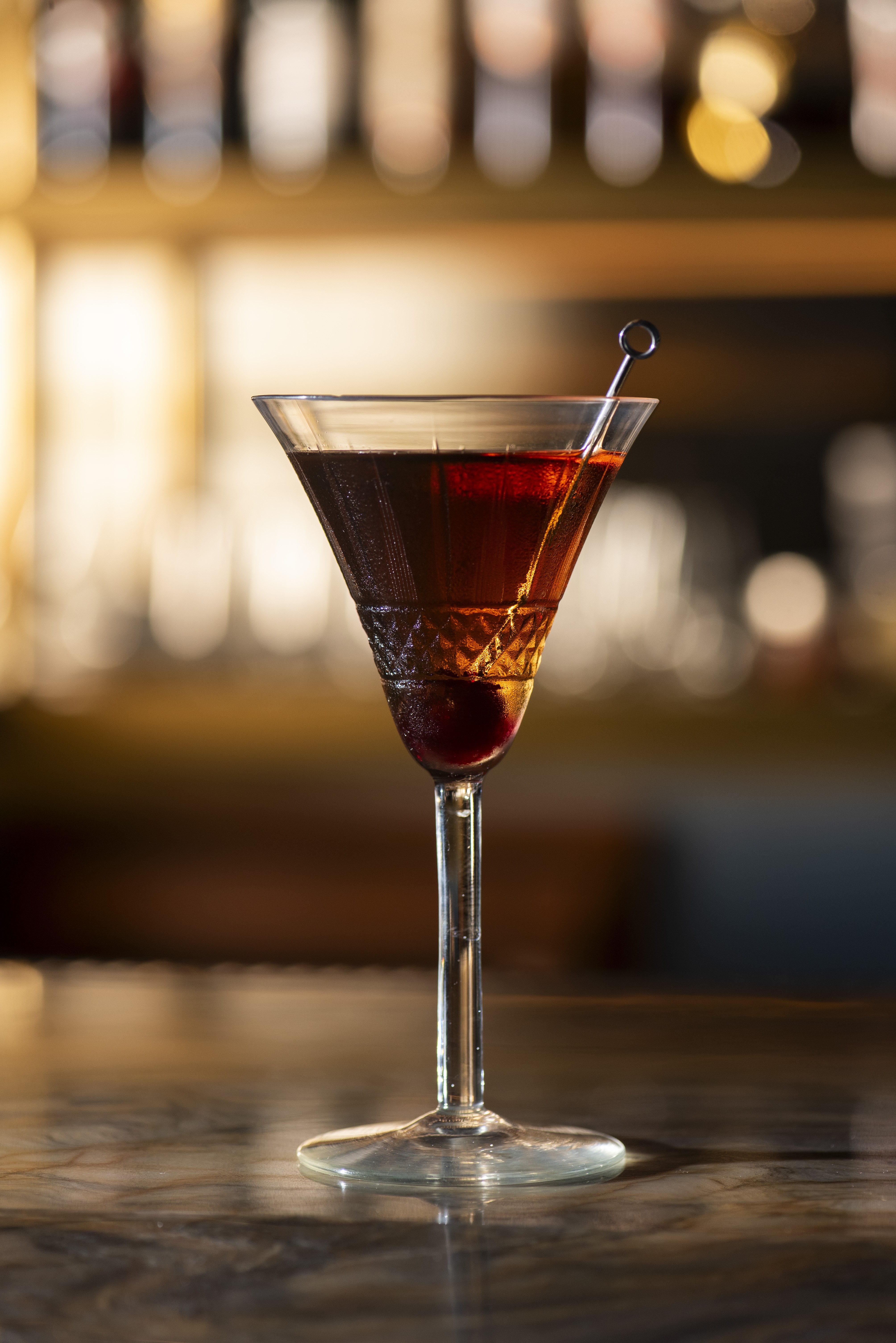 Coquetel vermelho escuro em ta&ccedil;a de martini com cereja no fundo e espeto met&aacute;lico, sobre balc&atilde;o de m&aacute;rmore, com fundo desfocado de garrafas iluminadas
