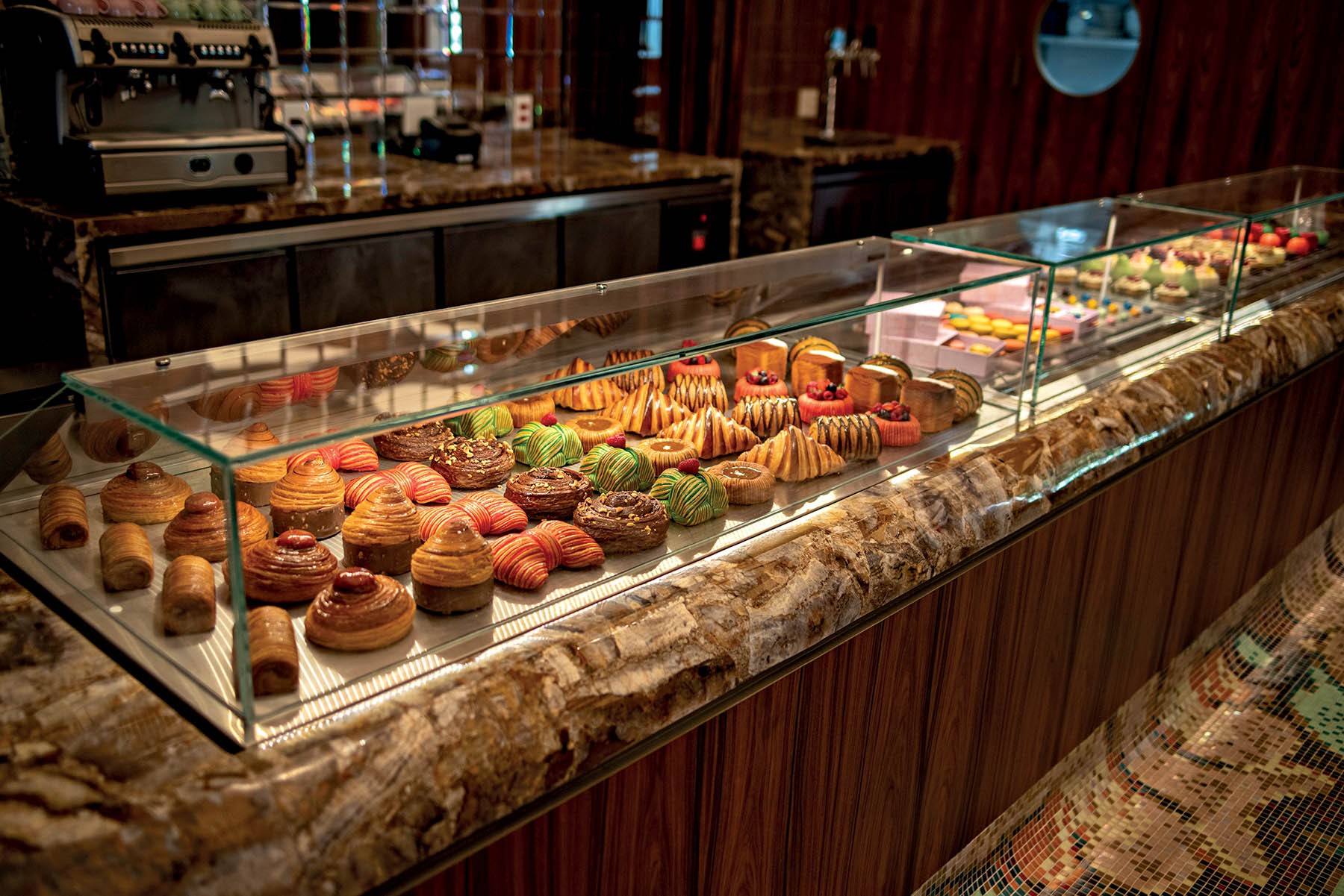 Exibição de doces e pães em uma vitrine de vidro, com uma variedade de croissants, bolos e tortas coloridas em tons de marrom, verde, vermelho e dourado. Ao fundo, uma máquina de café e um balcão escuro.