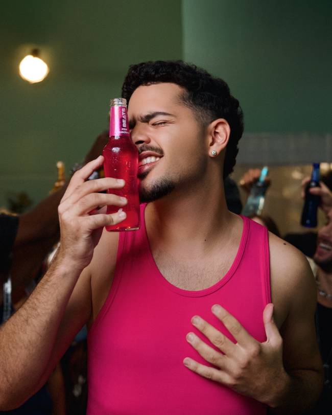 Homem jovem de pele morena, cabelo encaracolado e barba, vestindo regata rosa choque, lambe uma garrafa de Beats rosa com a l&iacute;ngua, os olhos fechados e a m&atilde;o no peito, em um ambiente de festa com luzes difusas
