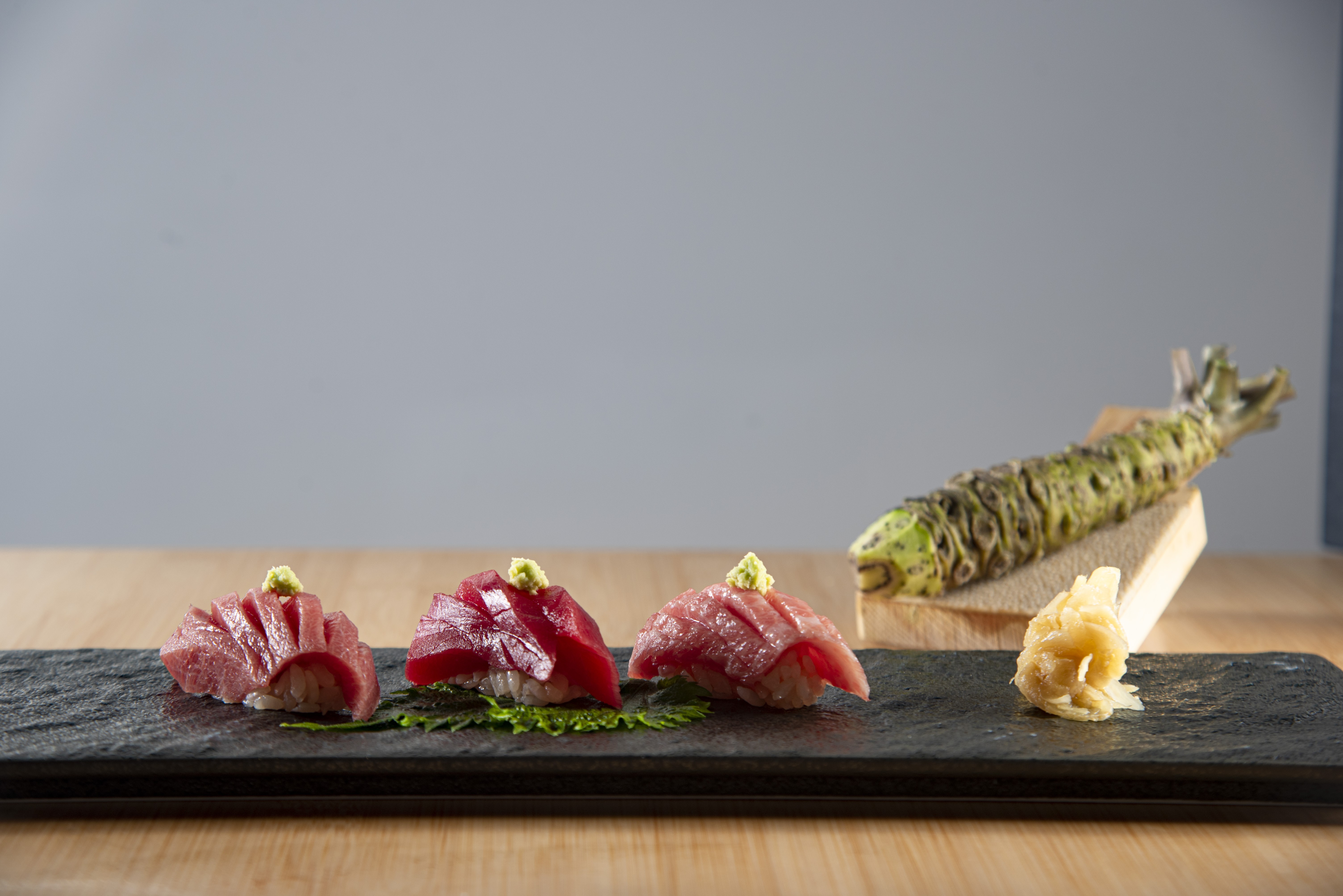 Tr&ecirc;s sushis de atum com wasabi no topo, sobre folhas verdes, dispostos em uma travessa preta de ard&oacute;sia. Ao lado, gengibre em conserva e um wasabi fresco em raiz sobre um suporte de madeira. O fundo &eacute; cinza claro e a superf&iacute;cie &eacute; de madeira clara