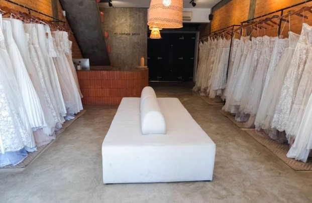 Interior de uma loja de vestidos de noiva com um sofá branco no centro. Fileiras de vestidos brancos pendurados em cabides ladeiam o ambiente. Ao fundo, uma escada e um balcão de tijolos. O teto tem luminárias de palha e spots pretos
