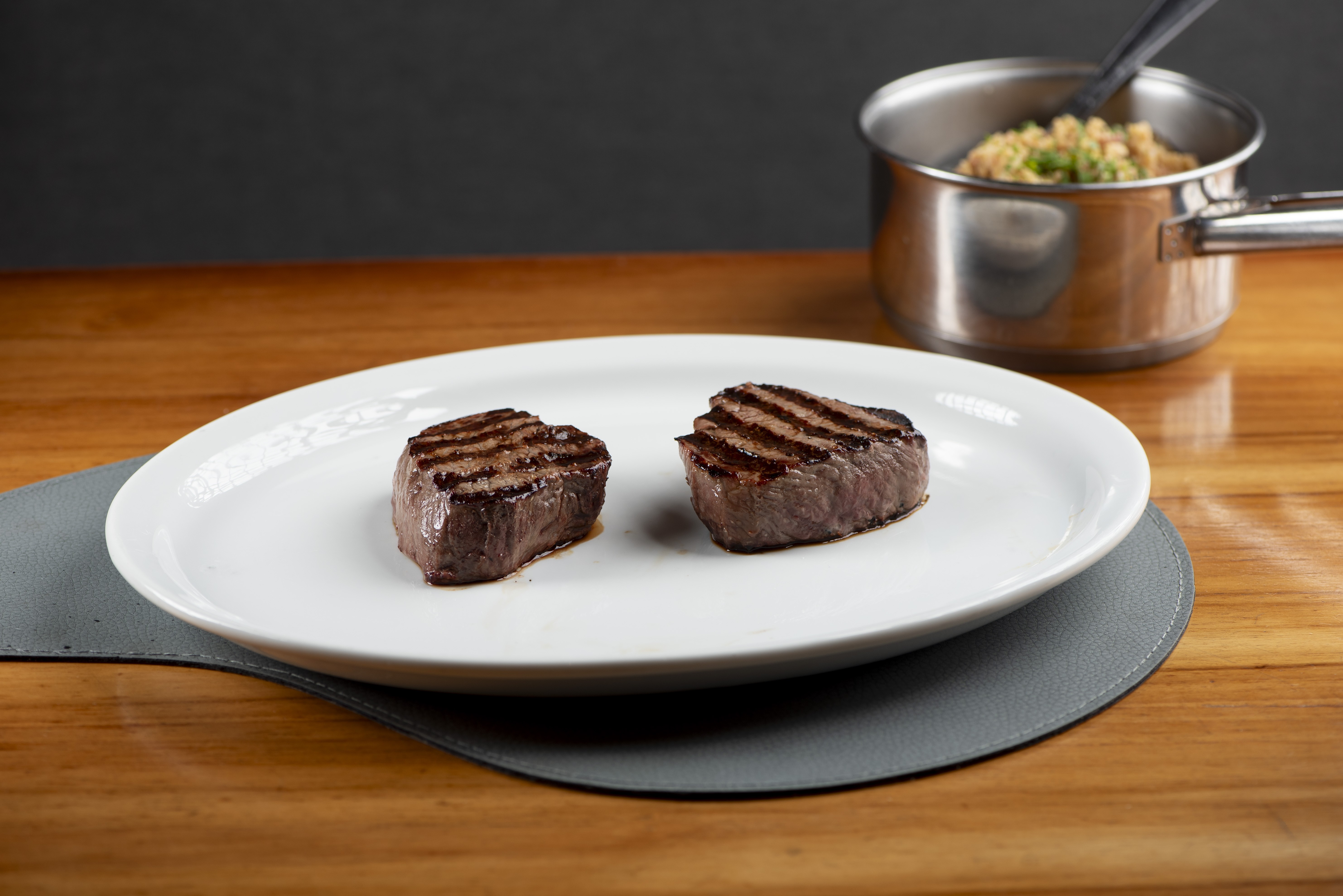 Dois medalh&otilde;es de carne grelhados com marcas de churrasco em um prato branco sobre um jogo americano cinza, em uma mesa de madeira. Ao fundo, uma panela de metal com arroz e cebolinha picada