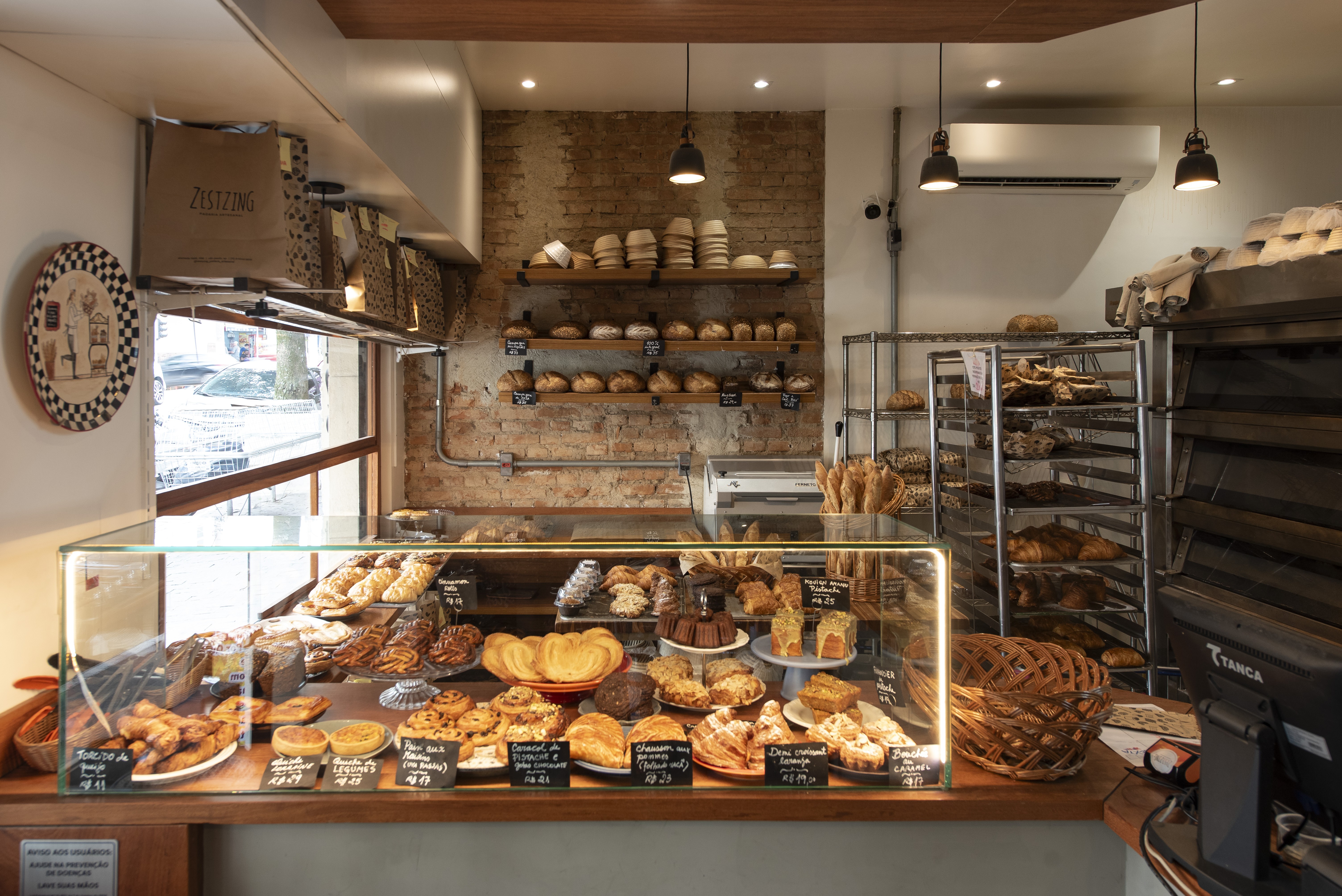 Interior de uma padaria com balc&atilde;o de vidro exibindo diversos doces e salgados, como croissants, tortas e p&atilde;es. Ao fundo, prateleiras de madeira com p&atilde;es variados e uma parede de tijolos aparentes. &Agrave; direita, estantes met&aacute;licas com assadeiras cheias de produtos de panifica&ccedil;&atilde;o e um forno industrial. Uma janela &agrave; esquerda mostra o exterior.