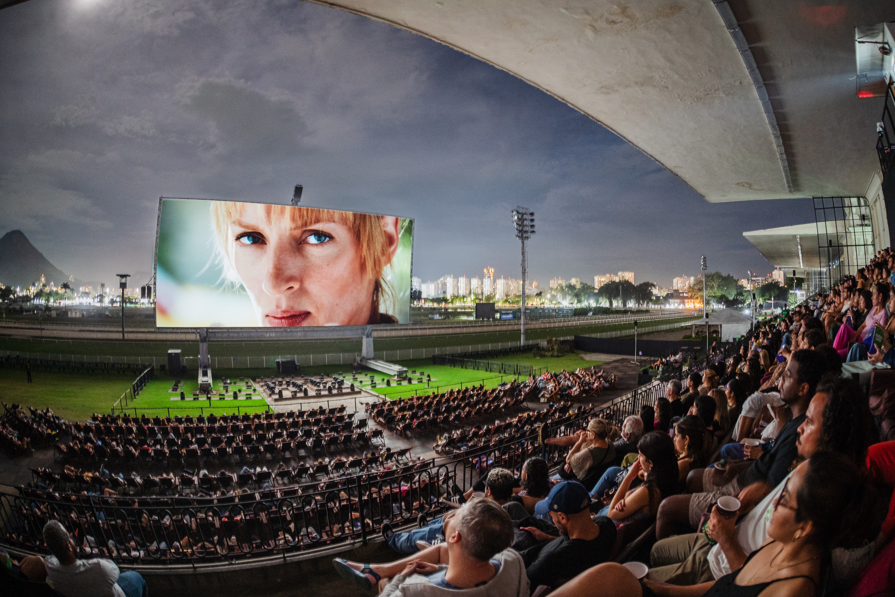 Pessoas assistem a um filme em um telão gigante ao ar livre, com a imagem de uma mulher loira de olhos azuis. O público está sentado em cadeiras dispostas em um gramado e em arquibancadas, sob um céu nublado. Ao fundo, a silhueta de montanhas e a cidade iluminada. Um projetor é visível no canto superior direito