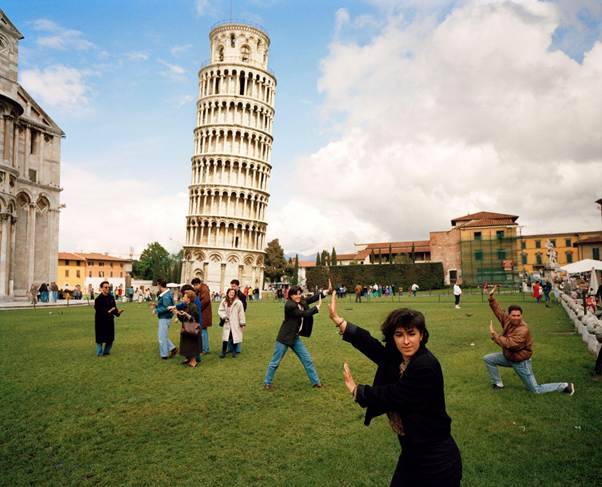 Turistas posam em frente &agrave; Torre de Pisa, na It&aacute;lia. Uma mulher em primeiro plano, de casaco preto, simula empurrar a torre com as m&atilde;os. Outros turistas fazem poses semelhantes no gramado, enquanto a torre inclinada se destaca sob um c&eacute;u parcialmente nublado
