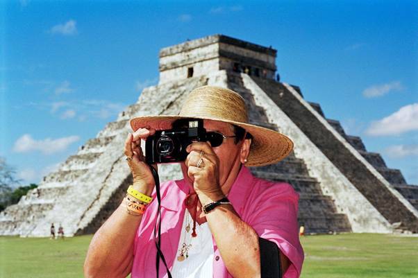 Mulher de chap&eacute;u de palha e &oacute;culos escuros, vestindo camisa rosa, fotografando com c&acirc;mera preta. Ao fundo, a pir&acirc;mide maia de Chich&eacute;n Itz&aacute; sob c&eacute;u azul com nuvens esparsas