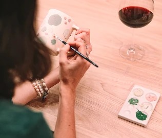 Pessoa pintando uma caneca de cerâmica branca com detalhes de patas de gato em tons pastel, usando um pincel fino. Na mesa de madeira clara, há uma taça de vinho tinto e uma paleta com tintas coloridas.