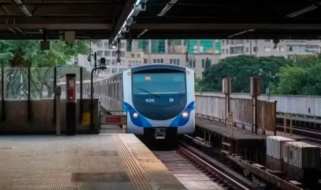 Um trem moderno branco e azul, modelo K25, se aproxima de uma plataforma de estação elevada. Ao fundo, prédios e árvores verdes compõem a paisagem urbana.
