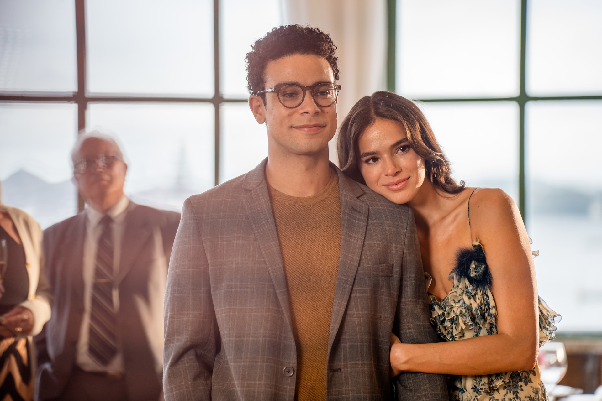 Um homem de óculos e blazer xadrez marrom claro sorri, enquanto uma mulher de vestido estampado floral e alças finas apoia a cabeça em seu ombro, sorrindo gentilmente. Ao fundo, desfocado, um homem de terno e gravata listrada e uma mulher com blusa clara.