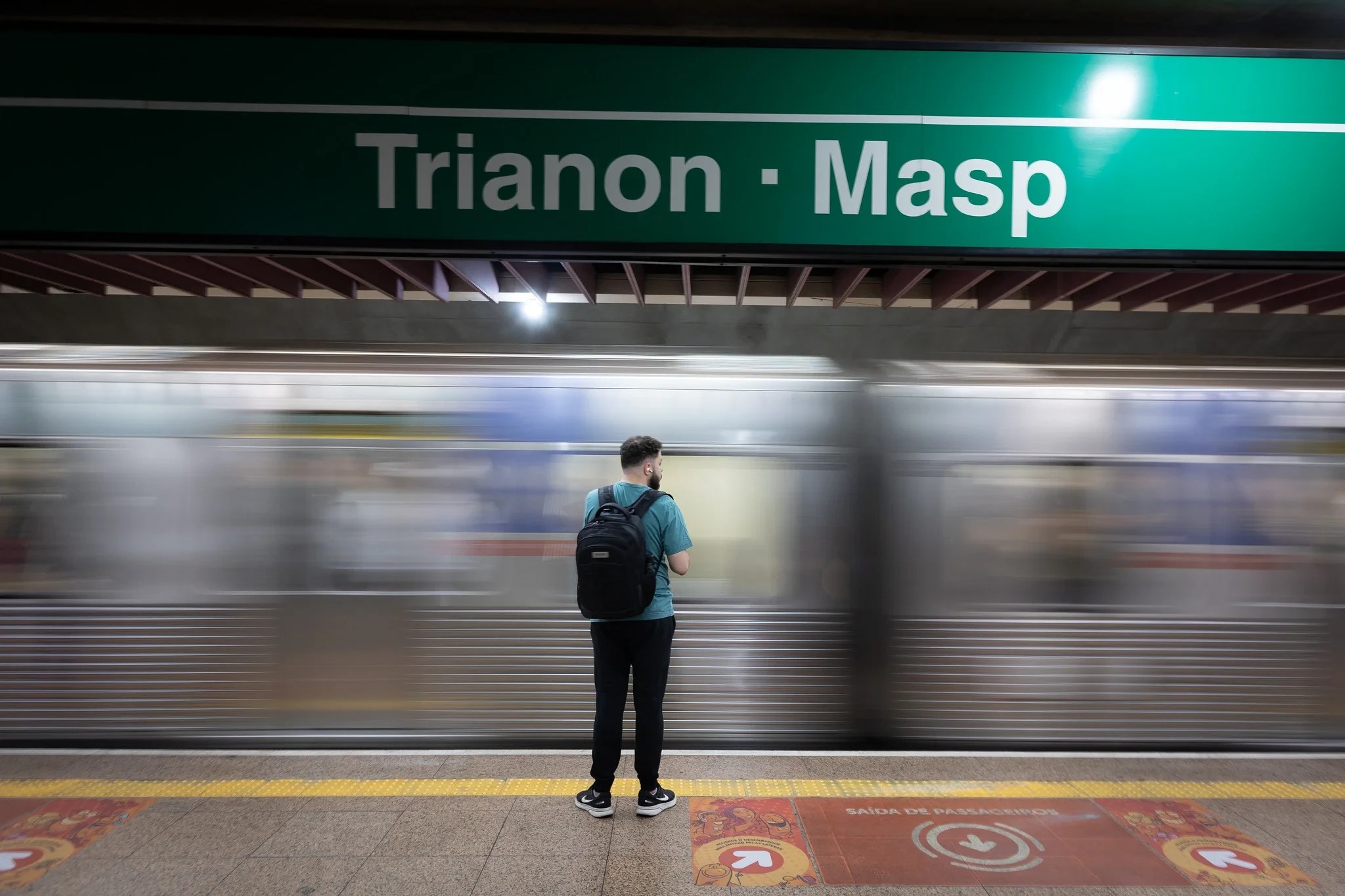 Homem de barba, camiseta azul e mochila preta, de costas, parado na plataforma da estação Trianon-Masp, enquanto um trem prateado passa em alta velocidade