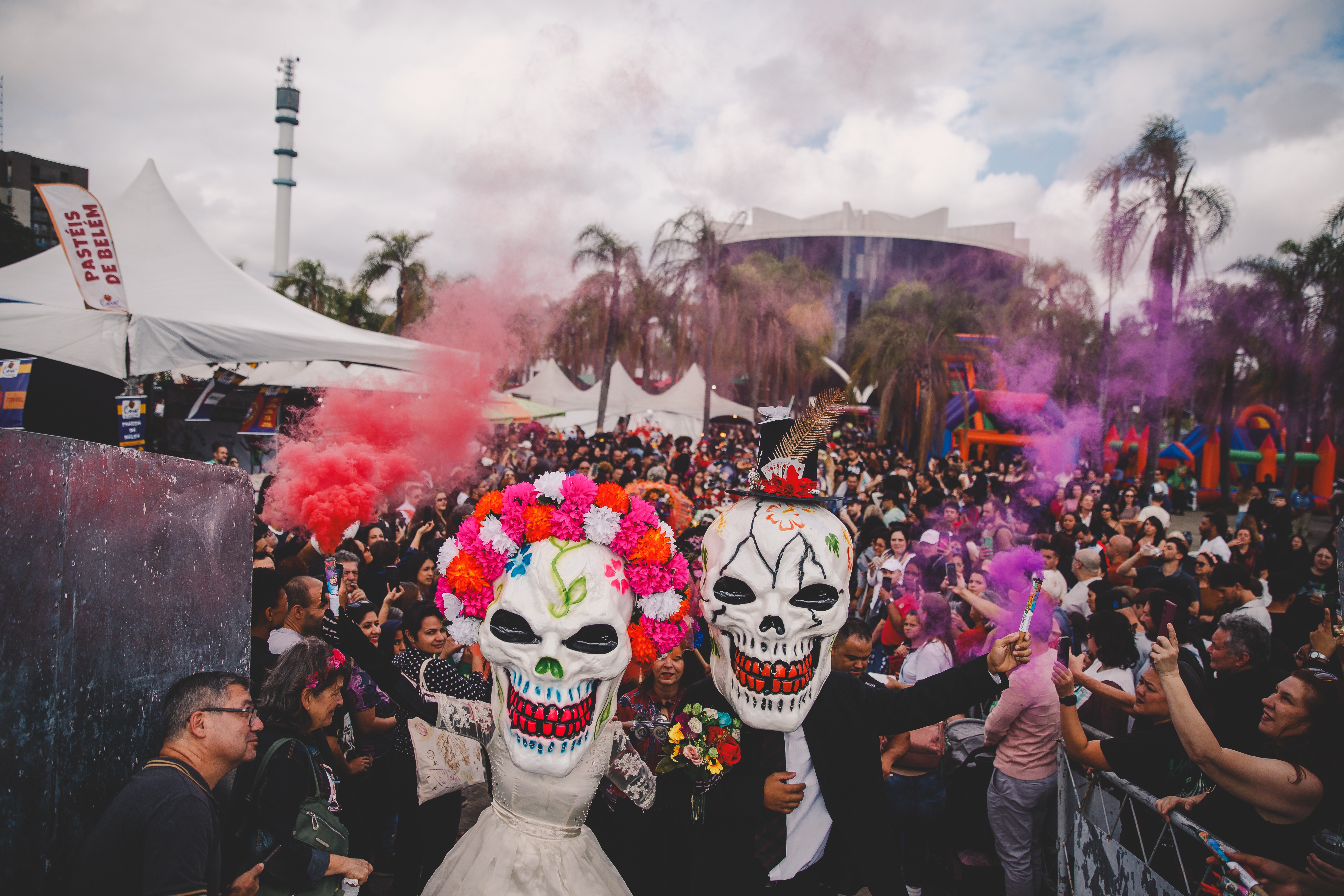 Duas pessoas fantasiadas de caveiras coloridas, uma com flores na cabeça e outra com cartola, posam em meio a uma multidão em festa. Fumaça rosa e roxa sobe no ar, com barracas brancas e uma torre ao fundo