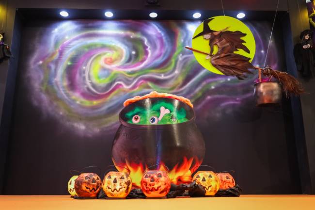 Decora&ccedil;&atilde;o de Halloween com caldeir&atilde;o preto borbulhante, olhos e um osso dentro, cercado por ab&oacute;boras luminosas. Ao fundo, uma proje&ccedil;&atilde;o de espiral colorida e uma bruxa voando em uma vassoura com um balde pendurado, em frente a uma lua amarela