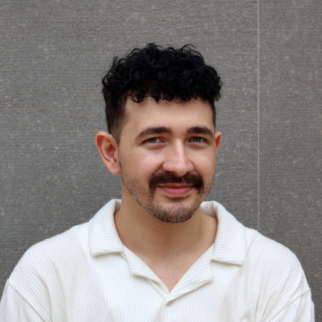 Homem jovem, pele clara, cabelo preto encaracolado e barba curta, bigode marcante, sorri levemente, veste camisa polo branca texturizada, fundo cinza liso