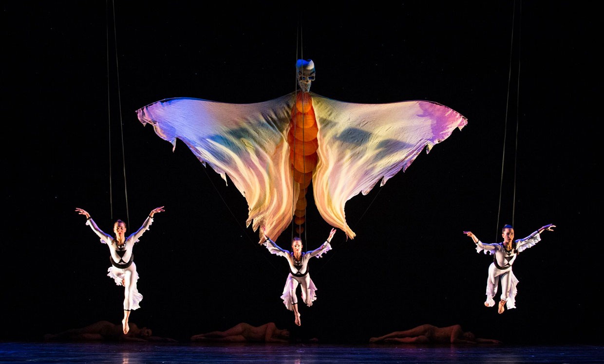 Três bailarinas em trajes brancos voam suspensas por cabos em um palco escuro, com os braços abertos. Ao centro, uma estrutura de tecido colorido, semelhante a uma borboleta gigante, paira sobre elas. No chão, figuras deitadas em tons terrosos.