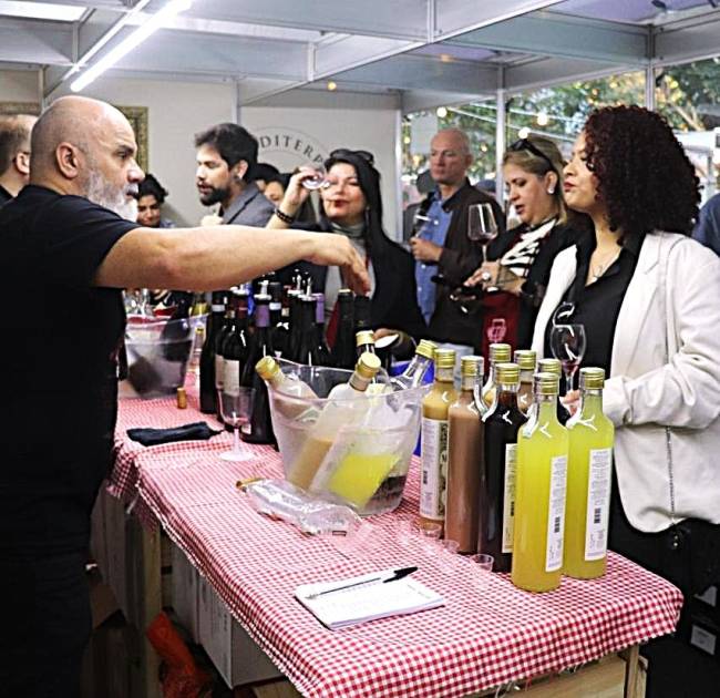 Homem careca com barba grisalha serve bebida em evento, enquanto pessoas provam vinhos e licores em um balc&atilde;o com toalha xadrez vermelha e branca