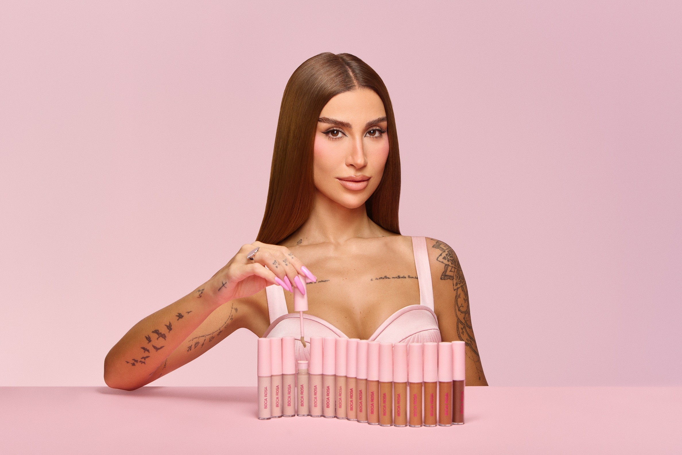 Mulher de cabelos castanhos e lisos, com maquiagem e unhas longas rosa, segura um aplicador de gloss labial. Ela veste um top rosa claro e exibe tatuagens nos braços e peito. À sua frente, uma fileira de 15 tubos de gloss em tons variados de nude e marrom, sobre uma superfície rosa. O fundo é rosa liso