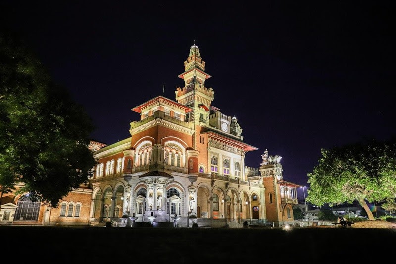 Palácio da Liberdade em Belo Horizonte à noite, iluminado, com arquitetura eclética em tons de terracota e branco, torre central e árvores frondosas nas laterais, sob céu escuro