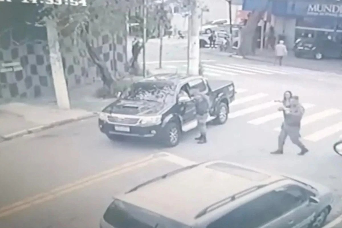 Câmera de segurança mostra dois homens armados abordando uma caminhonete preta em uma rua movimentada, um deles apontando a arma para o motorista, enquanto outro homem se aproxima da porta do passageiro. Ao fundo, pedestres e carros circulam normalmente, e um prédio com a palavra MUNDI é visível. A imagem está em preto e branco e com baixa resolução